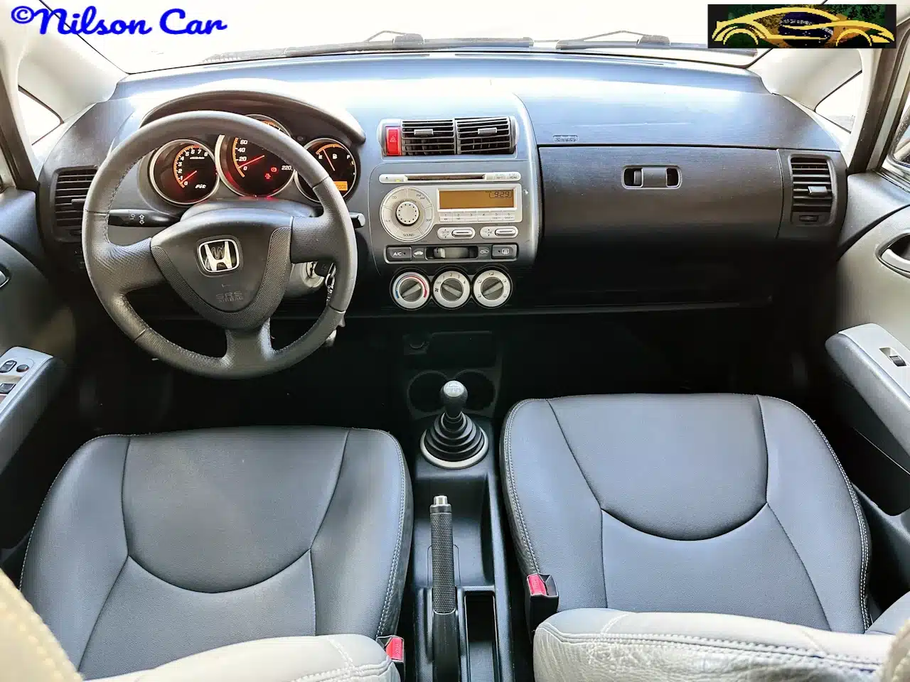 2008 Honda Fit EX 15 Prata - dash-WM - Nilson Car Interior view of a compact car showing the steering wheel, central dashboard with radio and climate controls, and a manual gear shift between two grey front seats. Watermarks read 'Nilson Car' and a small graphic logo are visible.