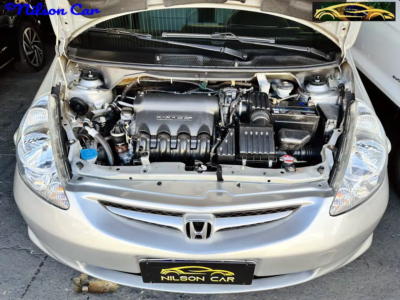 2008 Honda Fit EX 15 Prata - engine-WM - Nilson Car Front view of a silver Honda with the hood open, exposing the engine bay and components like battery and air intake; Nilson Car branding is visible on the license plate and top corner.
