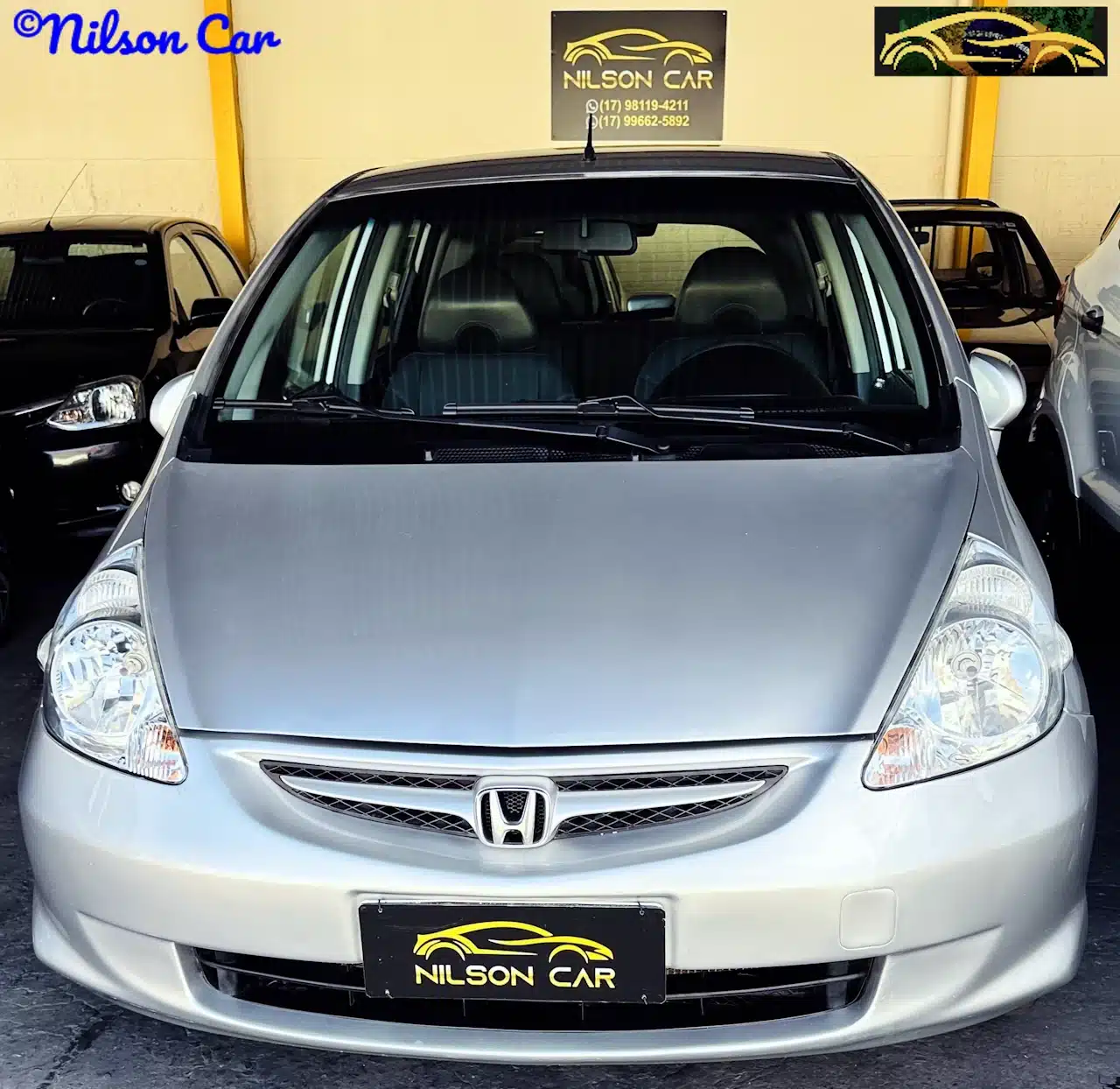 2008 Honda Fit EX 15 Prata - front-WM - Nilson Car Front view of a silver Honda hatchback at a car dealership, with a Nilson Car license plate sign visible on the bumper.