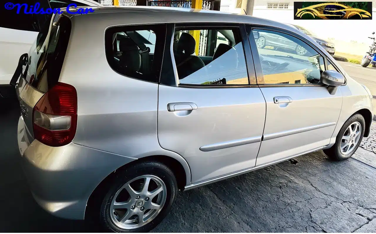 2008 Honda Fit EX 15 Prata - side2-WM - Nilson Car Silver hatchback car photographed from the rear-left, parked on a cracked pavement, with a 'Nilson Car' watermark in the top-left corner.