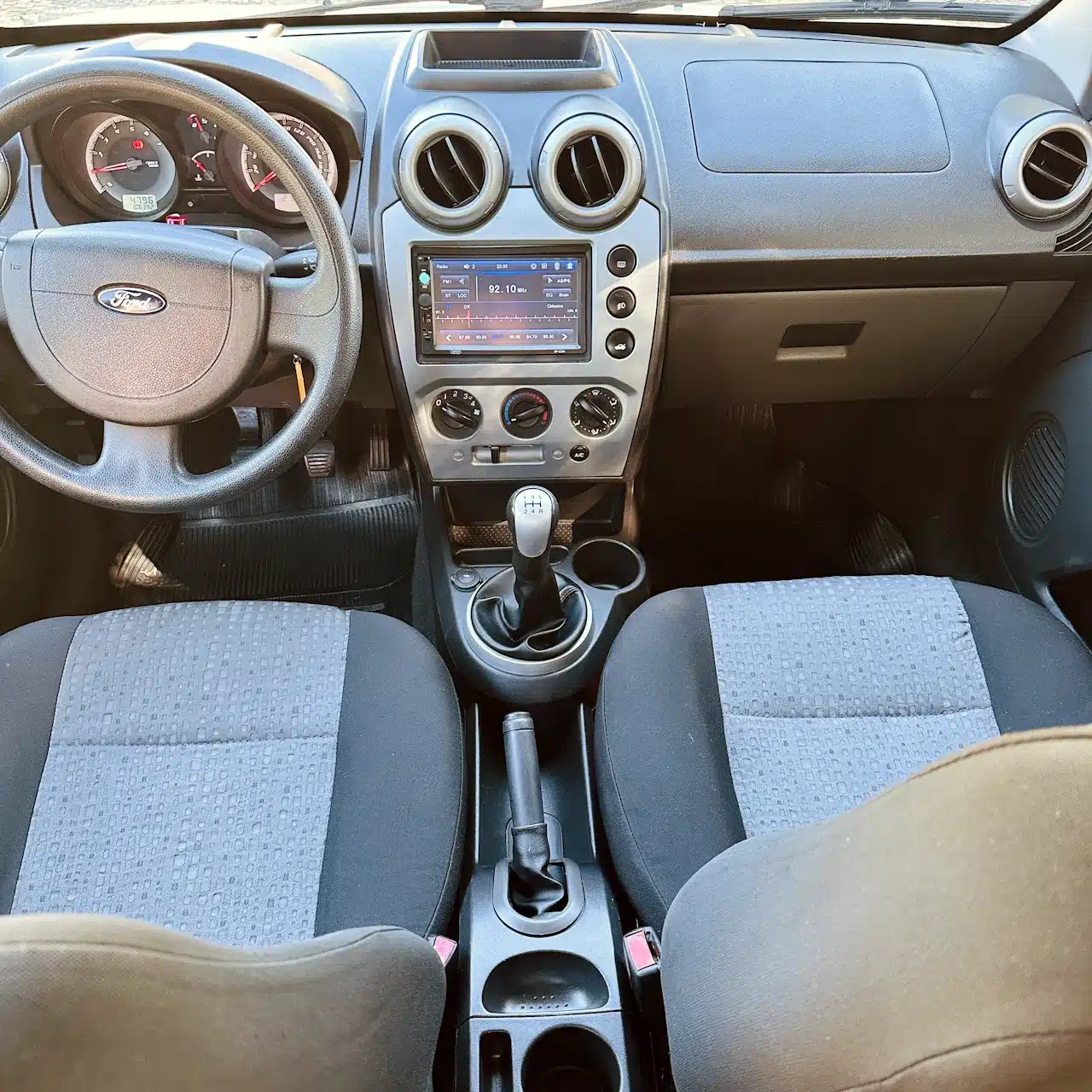 2013 Ford Fiesta Sedan 16 M Flex Branco - dash - Nilson Car Interior of a car cockpit showing steering wheel, gauge cluster, center touchscreen, round air vents, climate controls, and a manual gear shifter between front seats.