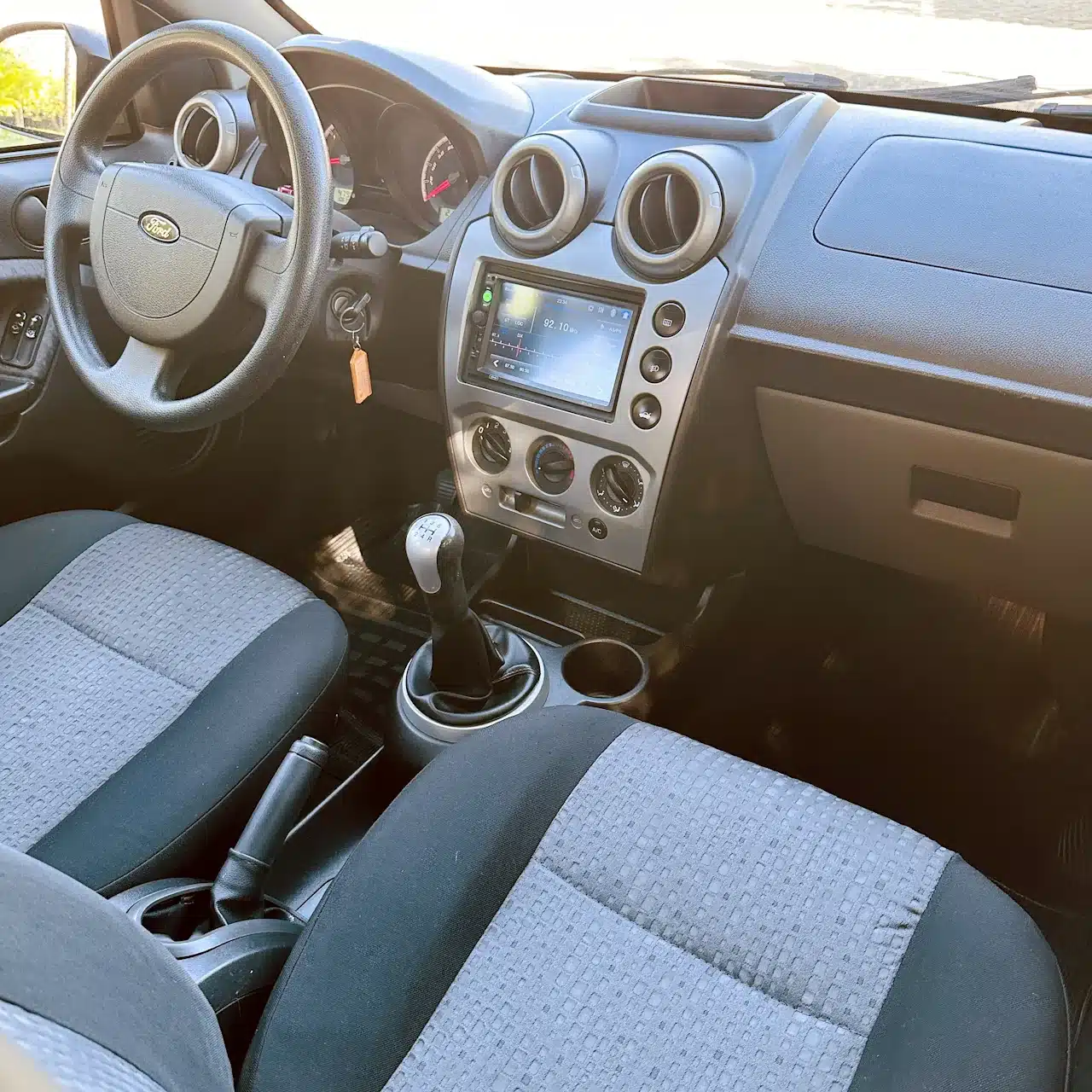 2013 Ford Fiesta Sedan 16 M Flex Branco - front-seat - Nilson Car Interior of a car focusing on the driver area: steering wheel, center console, and manual gear shift visible against gray seats.