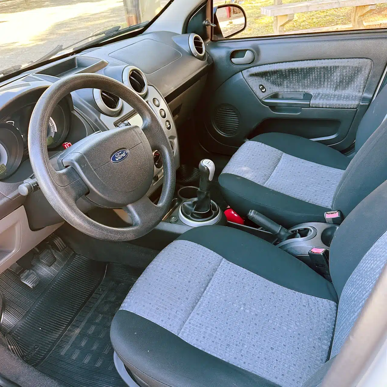2013 Ford Fiesta Sedan 16 M Flex Branco - front-seat2 - Nilson Car Interior view of a Ford car showing the steering wheel, manual gear stick, and gray fabric seats from the passenger side door open at daylight