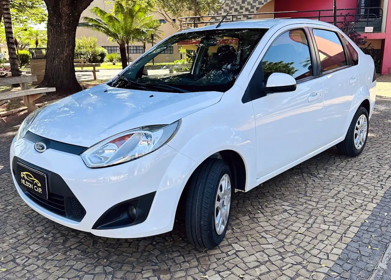 2013 Ford Fiesta Sedan 16 M Flex Branco - front-side - Nilson Car White Ford sedan parked on a cobblestone lot with trees in the background; roof rack installed and dealership plate visible on front