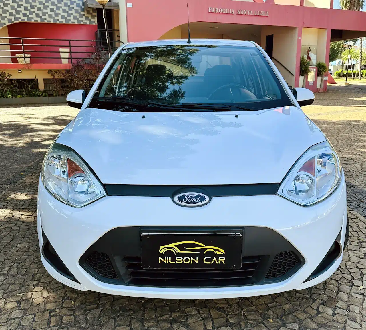 2013 Ford Fiesta Sedan 16 M Flex Branco - front - Nilson Car Front view of a white Ford hatchback parked on a cobblestone driveway; dealership plate reads NILSON CAR.