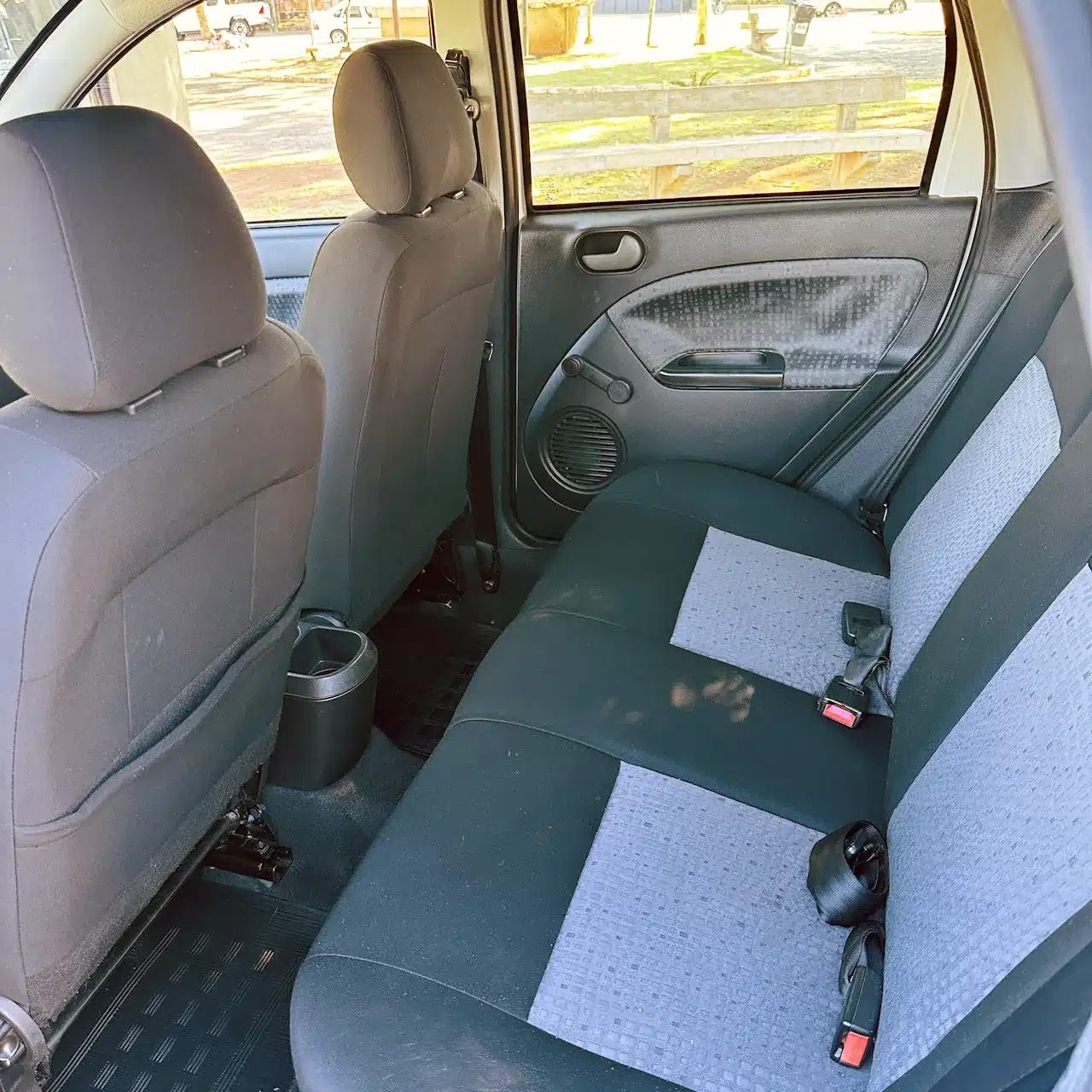 2013 Ford Fiesta Sedan 16 M Flex Branco - rear-seats - Nilson Car Back seat area of a small car with gray-blue upholstery and seat belts, view toward the rear door and window.