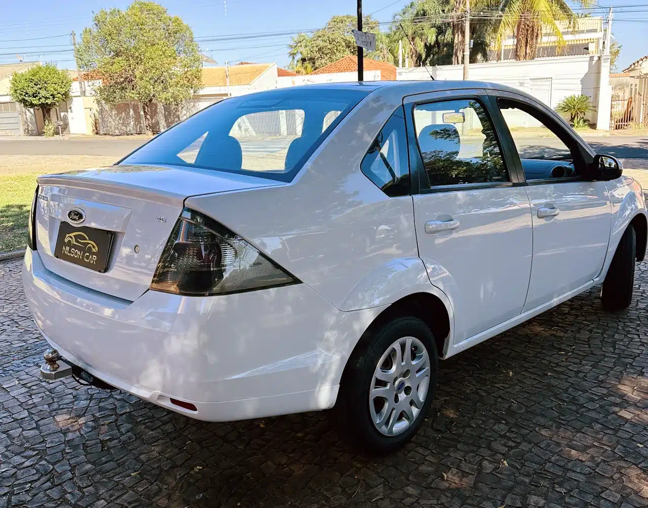 2013 Ford Fiesta Sedan 16 M Flex Branco - rear-side - Nilson Car White Ford sedan seen from rear-left, parked on a cobblestone driveway with a tow hitch visible beneath the bumper.