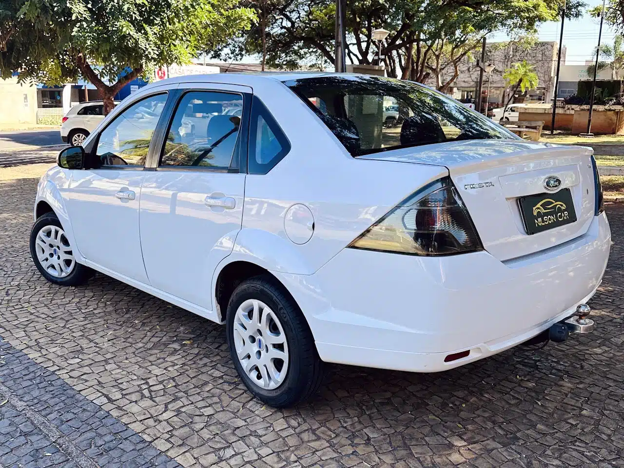 2013 Ford Fiesta Sedan 16 M Flex Branco - rear-side2 - Nilson Car White four-door sedan parked on a cobblestone lot, rear tow hitch visible and trees in the background.