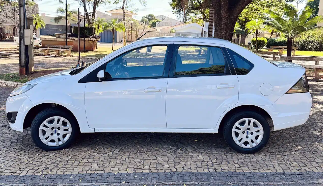 2013 Ford Fiesta Sedan 16 M Flex Branco - side - Nilson Car Side view of a white four-door sedan parked on a cobblestone street in a sunny park with trees and benches nearby.