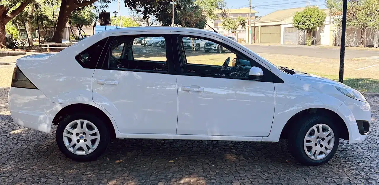 2013 Ford Fiesta Sedan 16 M Flex Branco - side2 - Nilson Car Side view of a white sedan parked on a cobblestone street during daytime, with trees and buildings in the background.