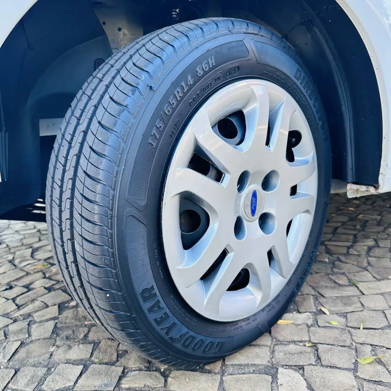 2013 Ford Fiesta Sedan 16 M Flex Branco - tire - Nilson Car Close-up of a car tire mounted on a silver Ford wheel with visible tread, on a paved surface