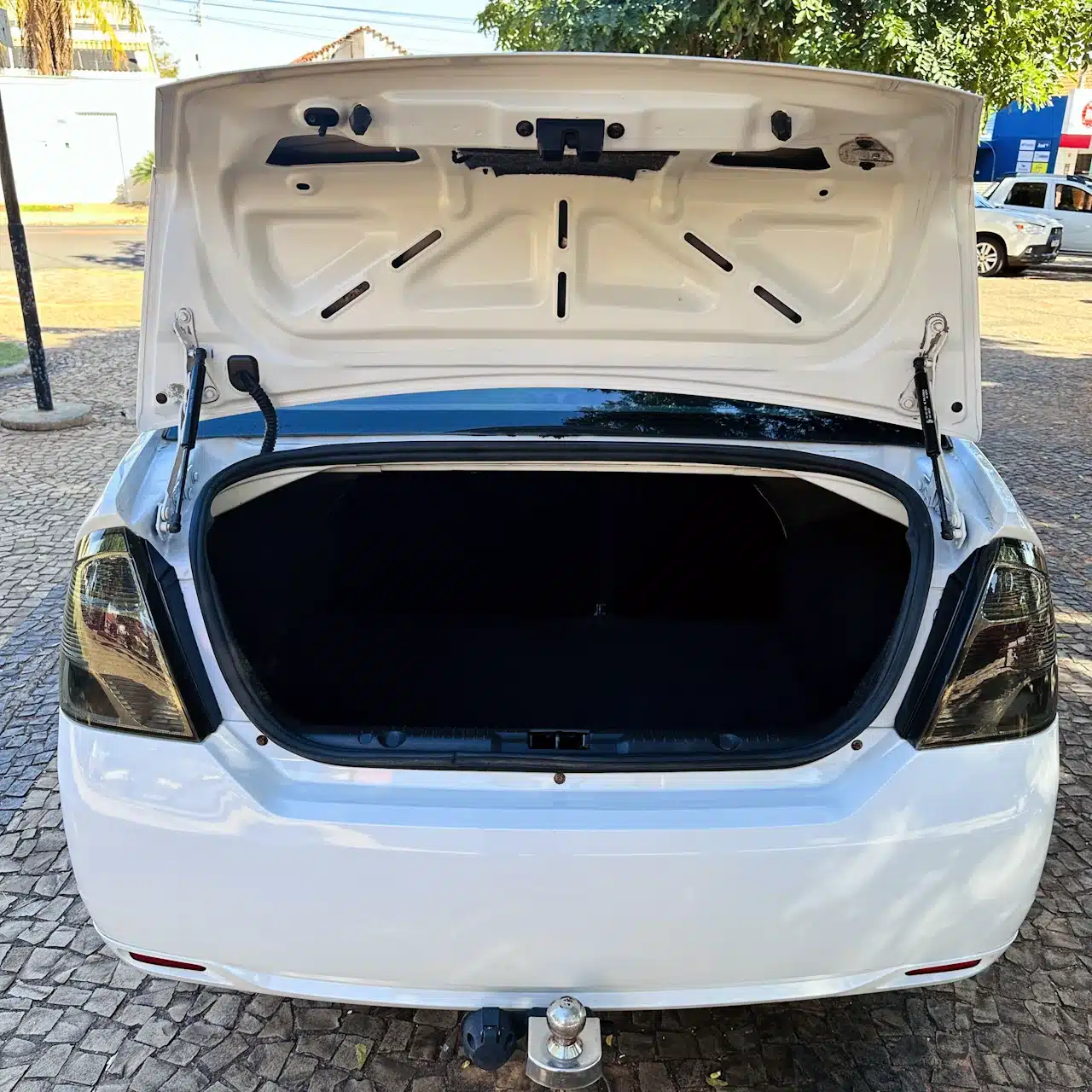 2013 Ford Fiesta Sedan 16 M Flex Branco - trunk - Nilson Car Open trunk of a white car showing a spacious empty cargo area, parked on a cobblestone street outside.