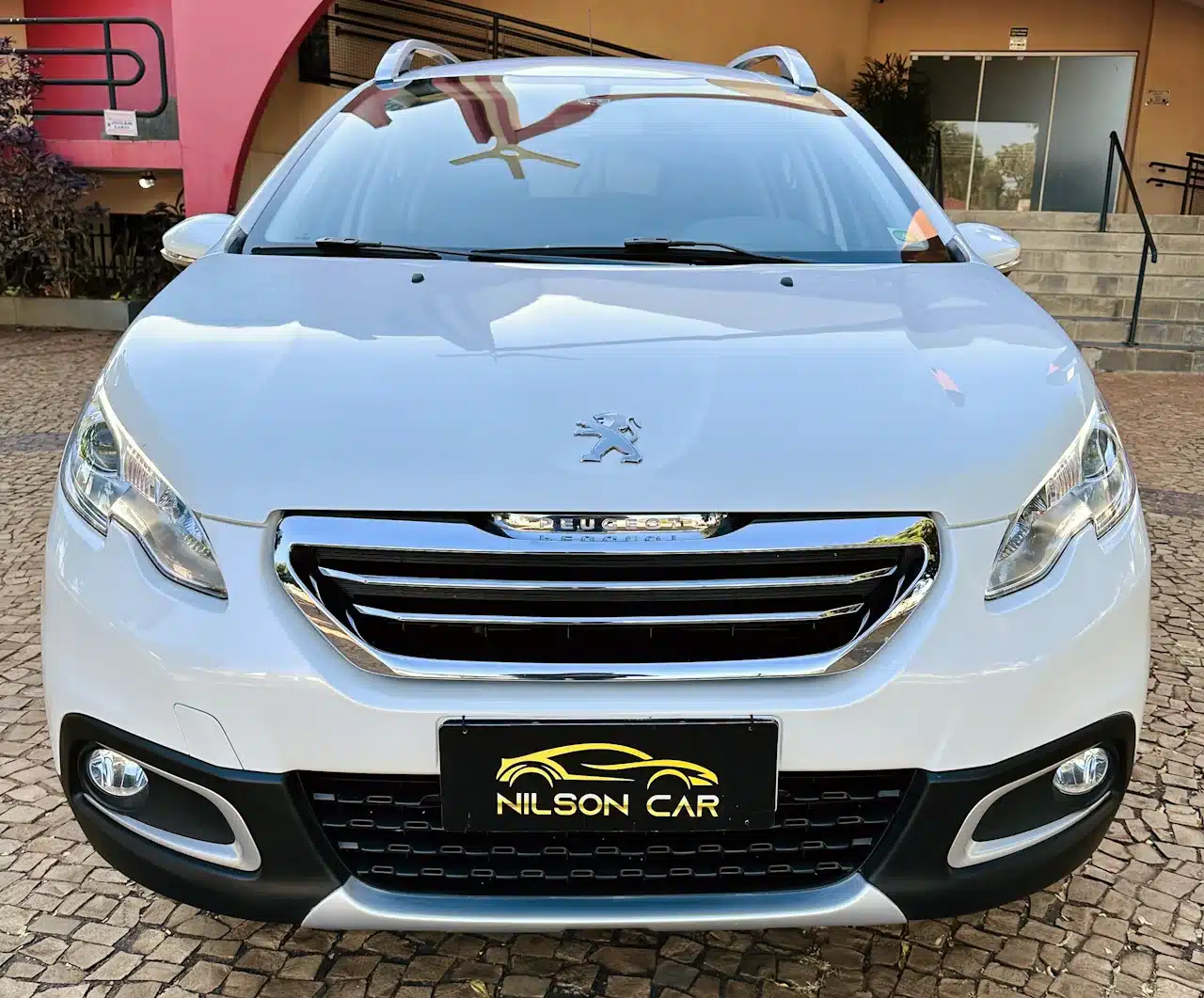 2016 PEUGEOT 2008 16 ALLURE BRANCA - front - Nilson Car Front view of a white Peugeot SUV with chrome grille and Nilson Car license plate on a cobblestone driveway.