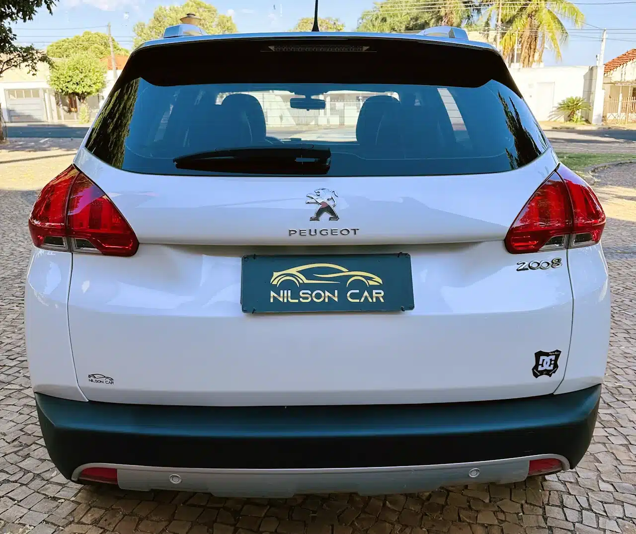 2016 PEUGEOT 2008 16 ALLURE BRANCA - rear - Nilson Car Rear view of a white Peugeot 2008 SUV, showing tail lights, emblem, and a teal license plate that reads NILSON CAR on a cobblestone driveway.
