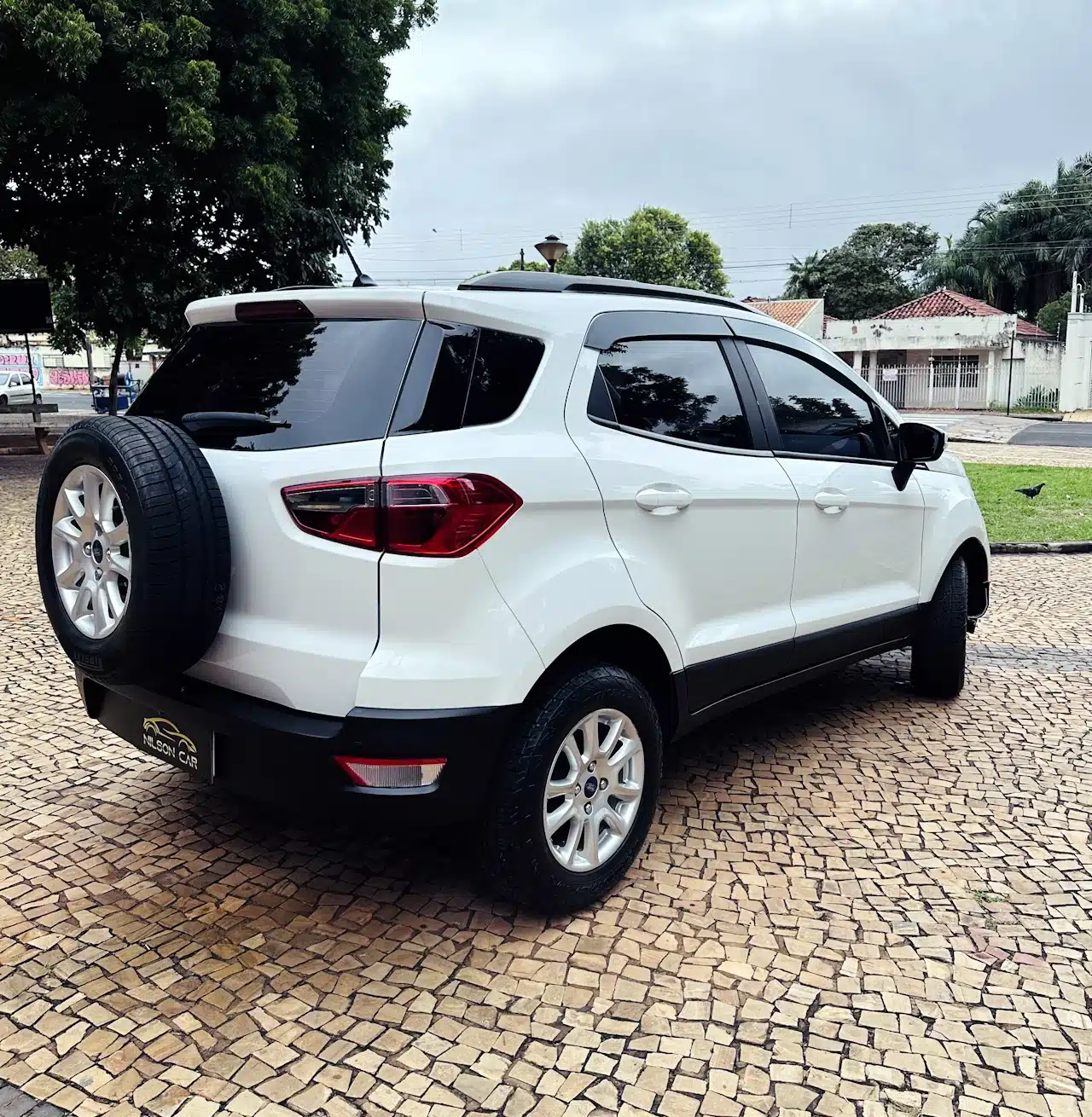 2020-21-Ford-Ecosport-SE-1_5-Branco-back-side - Nilson Car White SUV parked on a cobblestone driveway, with a spare tire mounted on the rear door and tinted rear windows.
