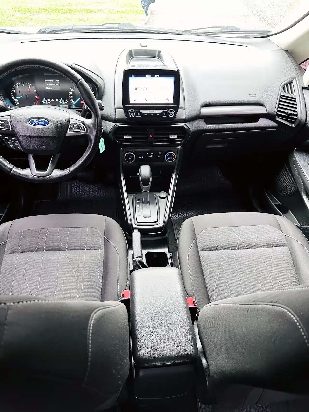 2020-21-Ford-Ecosport-SE-1_5-Branco-dash - Nilson Car Front interior of a Ford car showing steering wheel, center touchscreen, and gear shifter between two gray seats.