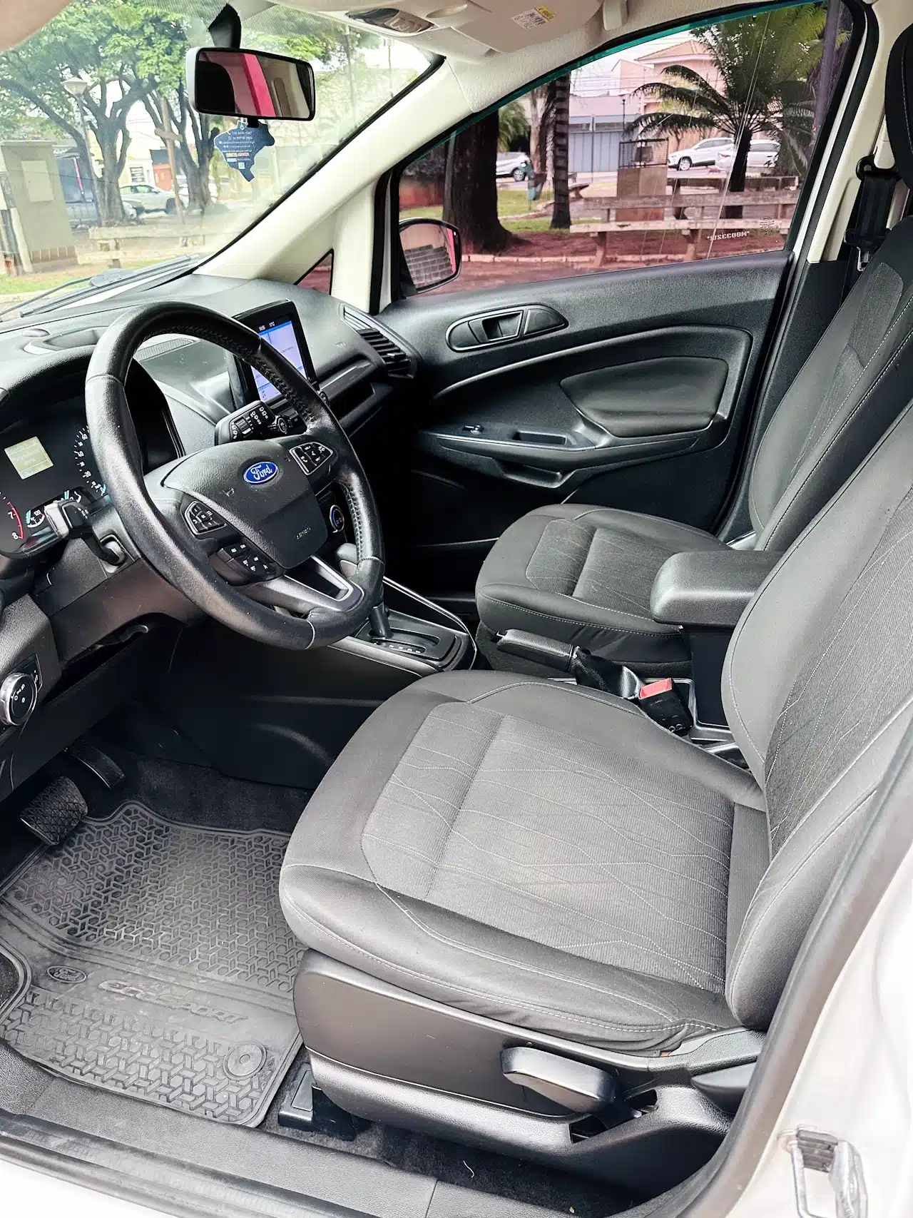 2020-21-Ford-Ecosport-SE-1_5-Branco-front-seats - Nilson Car Front interior view of a Ford car showing the steering wheel, dashboard screen, and grey fabric seats with rubber floor mats in the foreground.