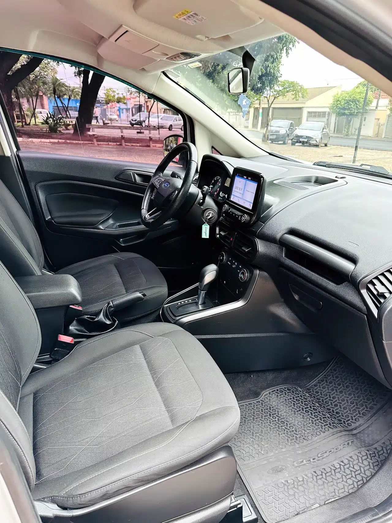 2020-21-Ford-Ecosport-SE-1_5-Branco-front-seats2 - Nilson Car Front interior of a car showing the steering wheel, center touchscreen, gear shifter, and two black fabric seats with a view of the street outside the windows.