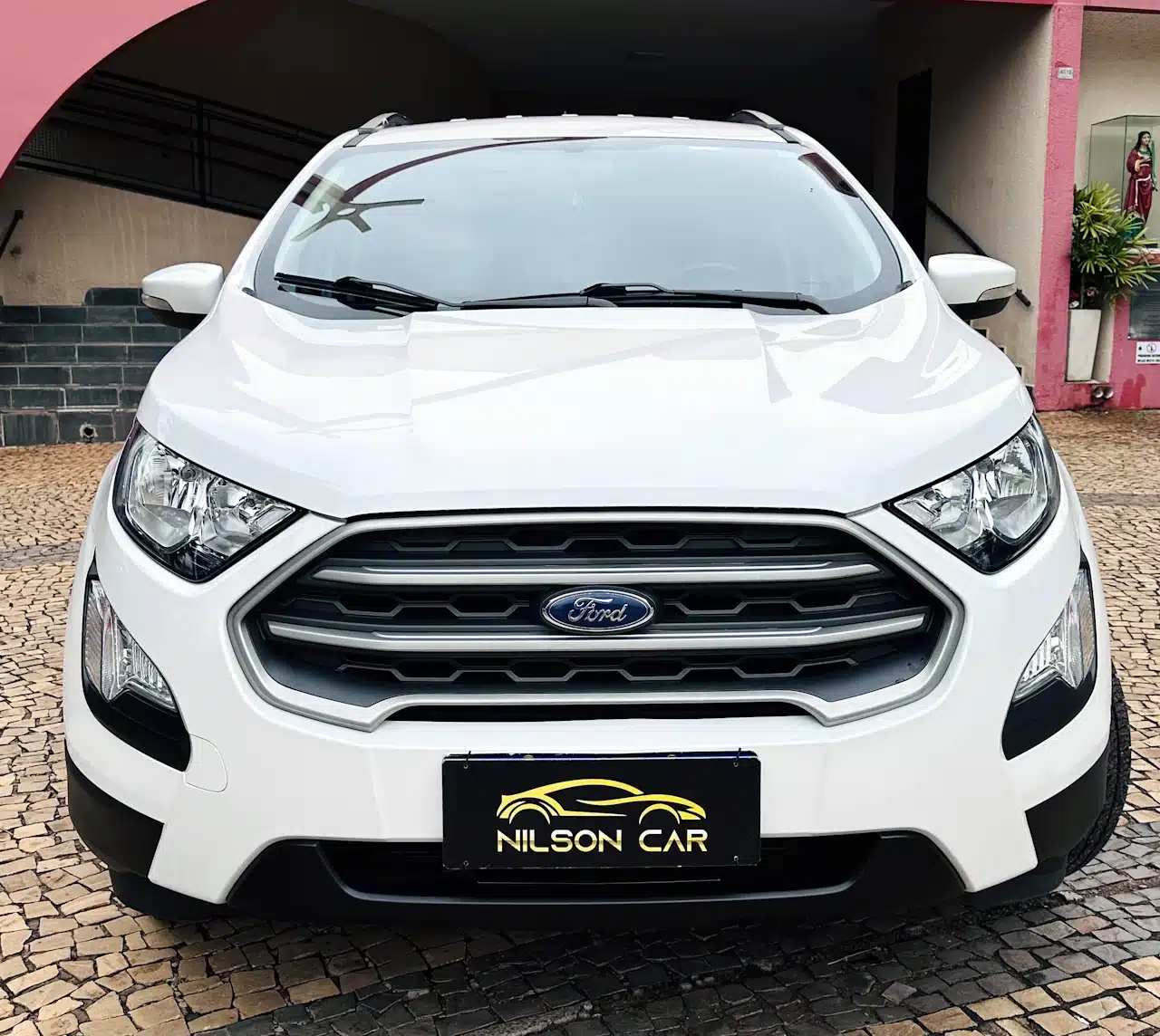 2020-21-Ford-Ecosport-SE-1_5-Branco-front - Nilson Car Front view of a white Ford SUV parked on a cobblestone driveway, with a Nilson Car license plate visible under the grille.