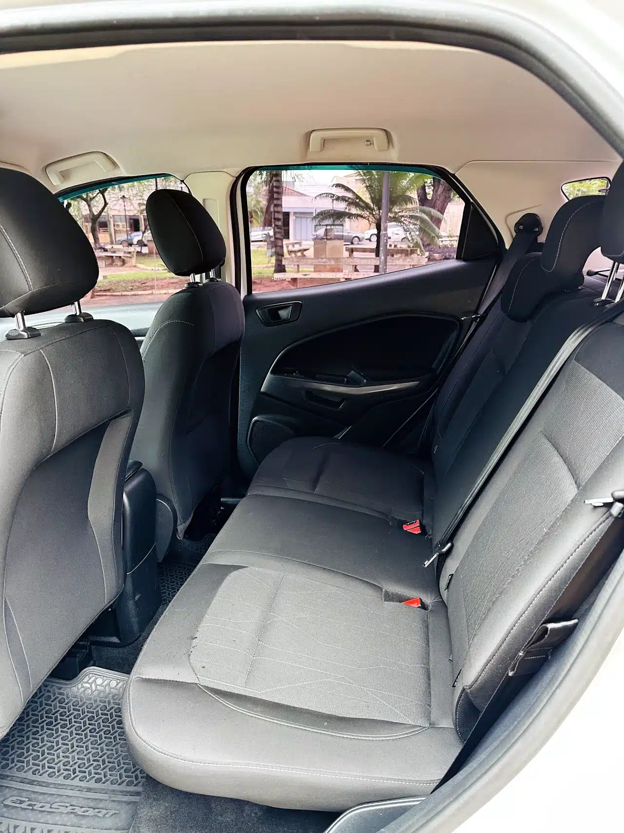 2020-21-Ford-Ecosport-SE-1_5-Branco-rear-seats - Nilson Car Rear seating area of a car with gray fabric seats and headrests, seen from the open door with a park and parked cars outside the window.