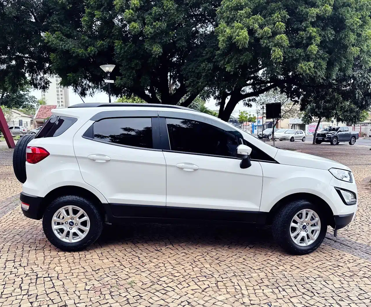 2020-21-Ford-Ecosport-SE-1_5-Branco-side - Nilson Car Side view of a white compact SUV parked on a patterned stone surface with trees in the background.