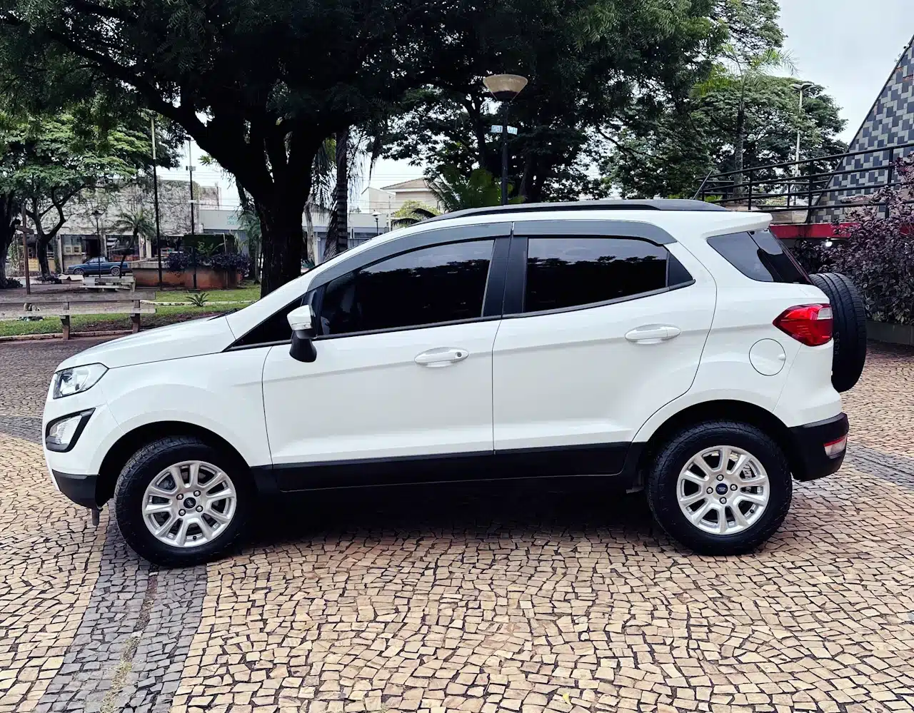 2020-21-Ford-Ecosport-SE-1_5-Branco-side2 - Nilson Car White compact SUV parked on a tan mosaic pavement with trees and buildings in the background (side view).