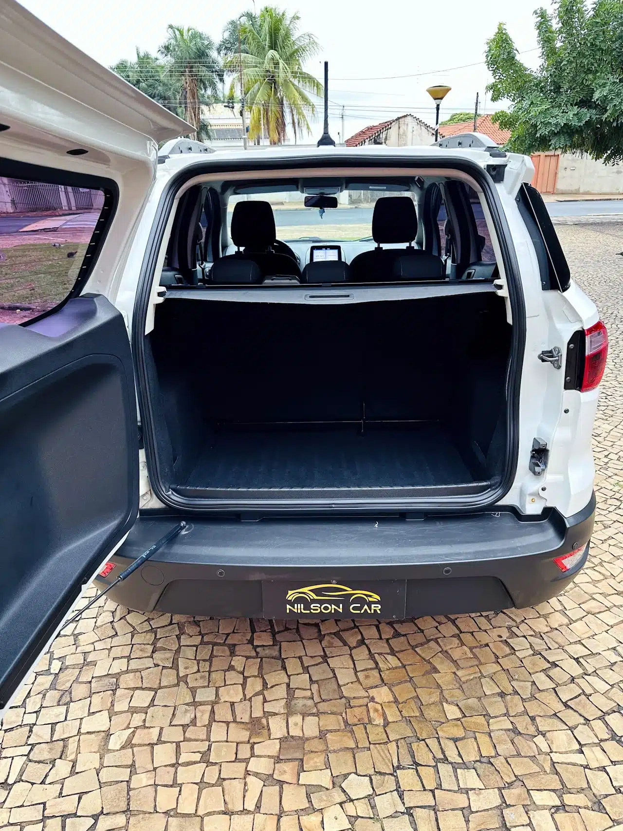 2020-21-Ford-Ecosport-SE-1_5-Branco-trunk - Nilson Car Open SUV cargo area with tailgate up; empty black luggage space, rear seats visible, Nilson Car logo on bumper.
