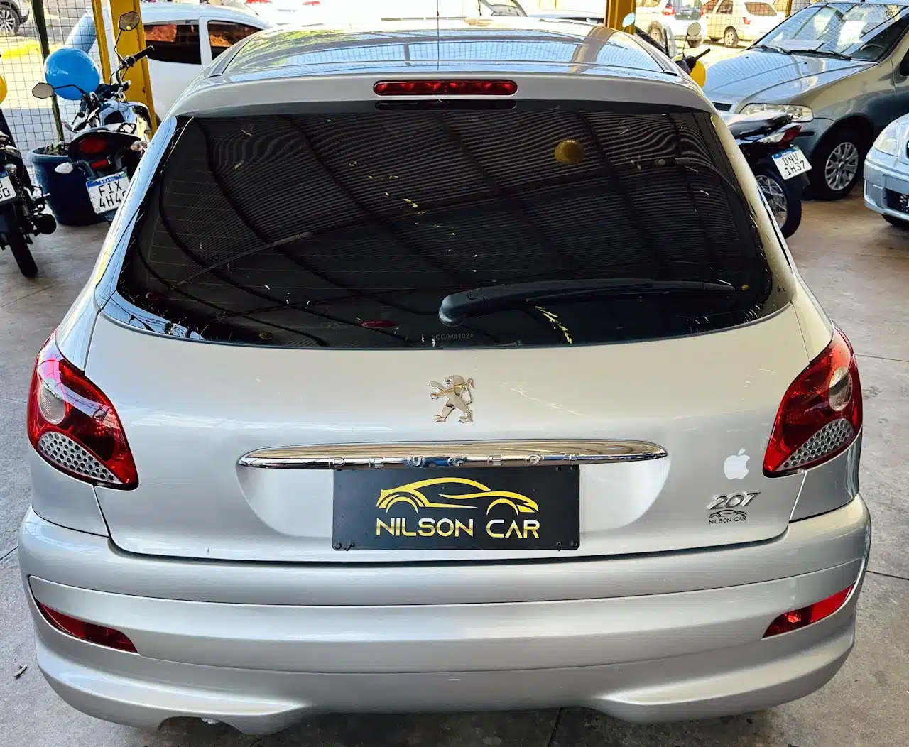Peugeot_ 207_ 2011_2012_Prata-back - Nilson Car Rear of a silver Peugeot 207 in a car dealership lot, with a NILSON CAR plate and an Apple logo sticker on the right trunk.