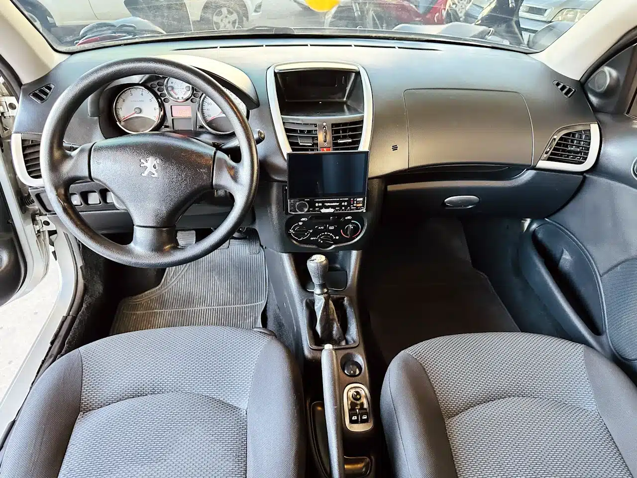 Peugeot_ 207_ 2011_2012_Prata-dash - Nilson Car Interior of a car cockpit facing the driver: steering wheel, instrument cluster, center touchscreen, manual gear stick, and front seats visible.