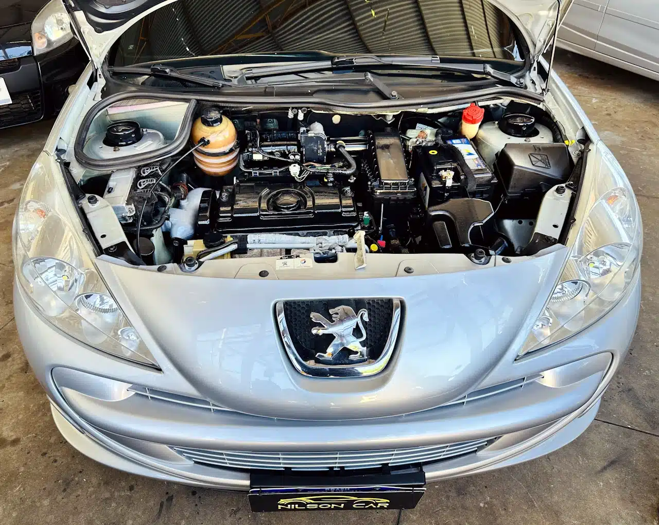 Peugeot_ 207_ 2011_2012_Prata-engine - Nilson Car Front view of a silver Peugeot with hood open, showing the car engine bay and surrounding components