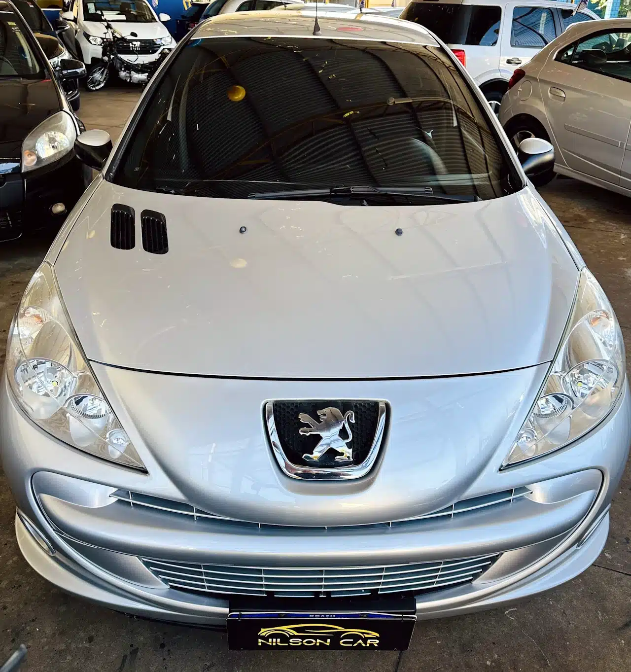 Peugeot_ 207_ 2011_2012_Prata-front - Nilson Car Front view of a silver Peugeot car in a showroom with its emblem visible on the grille and a Nilson Car plate.