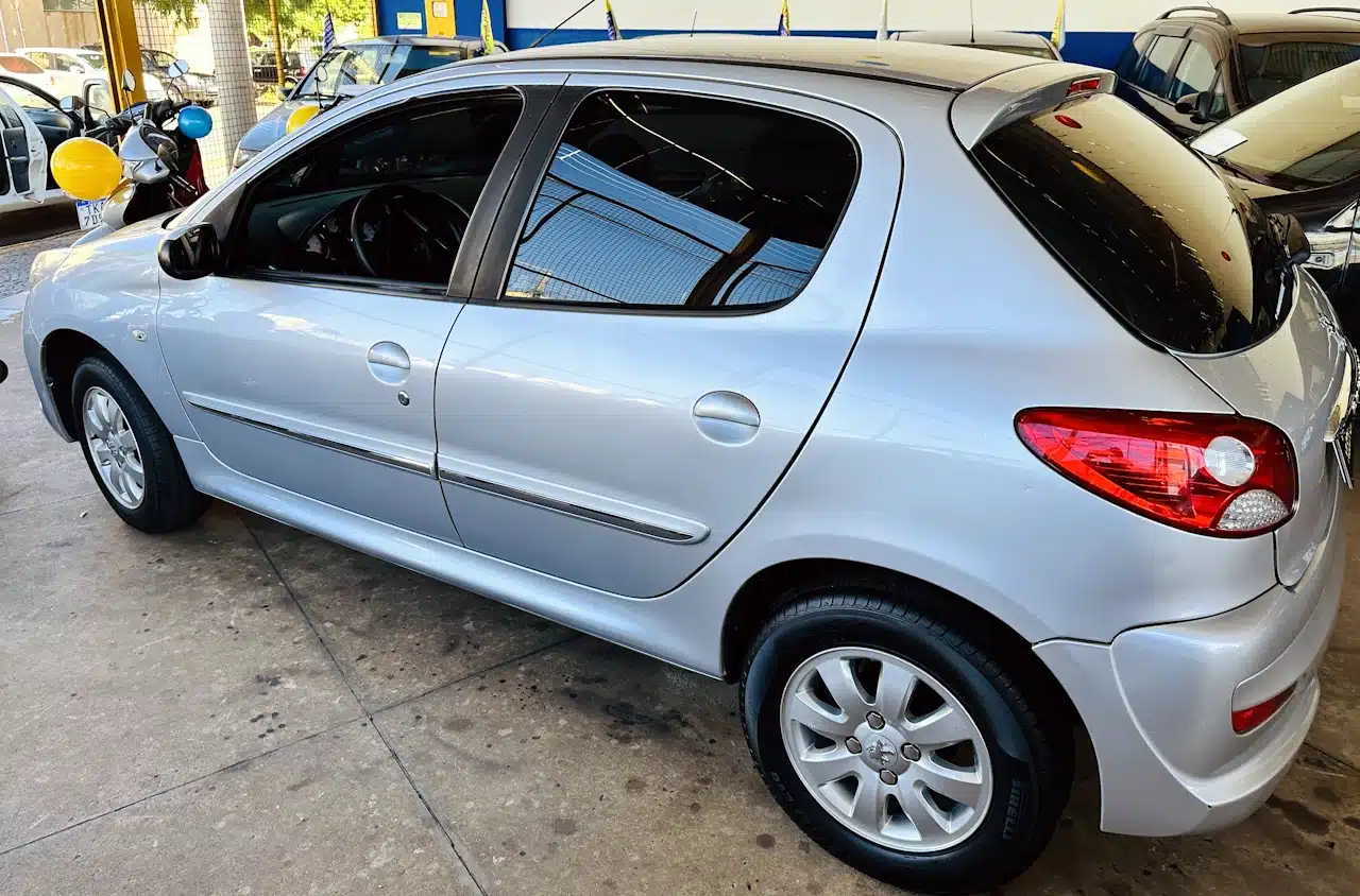 Peugeot_ 207_ 2011_2012_Prata-side - Nilson Car Silver hatchback car parked indoors at a dealership, showing the passenger side with rear and side doors visible.
