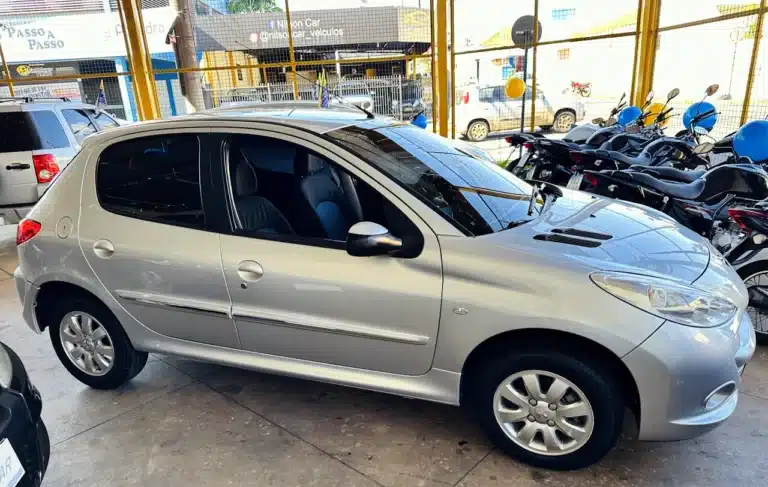 Peugeot_ 207_ 2011_2012_Prata-side2 - Nilson Car Three-quarter view of a silver compact hatchback parked inside a showroom, with motorcycles behind a yellow fence in the background.