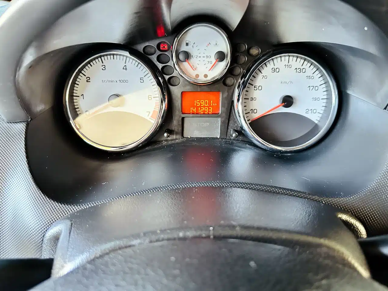 Peugeot_ 207_ 2011_2012_Prata-speedo - Nilson Car Dashboard instrument cluster with a left tachometer, right speedometer, and a central red digital display showing trip/odometer; steering wheel at bottom.